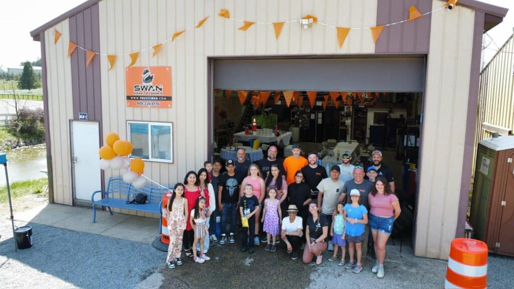 Group of employees and children standing in front of a Swan Co. garage decorated with balloons and flags.
