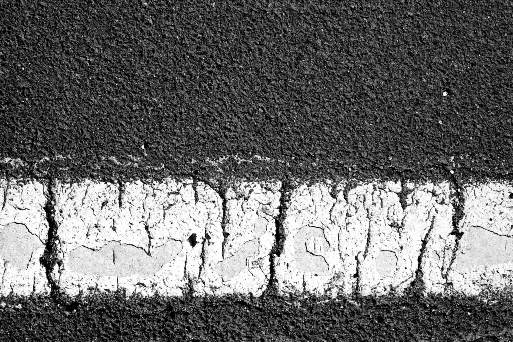 Close-up of sun-damaged asphalt with cracked white striping, showing signs of surface oxidation and binder aging. Example of summer pavement wear evaluated by Swan Co, an OSHA- and NAPA-certified paving contractor.