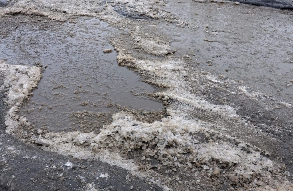 Pothole with standing water and slush showing late winter asphalt damage in a parking lot.