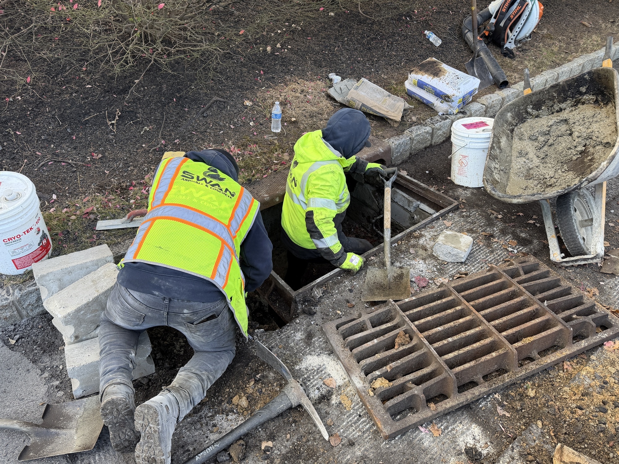 Workers repairing a catch basin drain in a parking lot to address drainage and water flow issues.